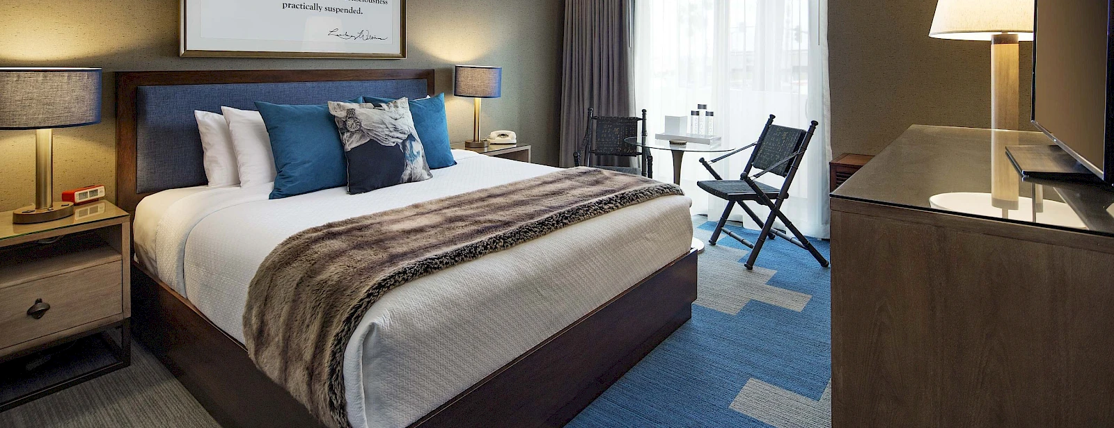 A modern hotel room with a plush king bed, dark wood furniture, blue accents, a seating area by the window, and soft lighting.