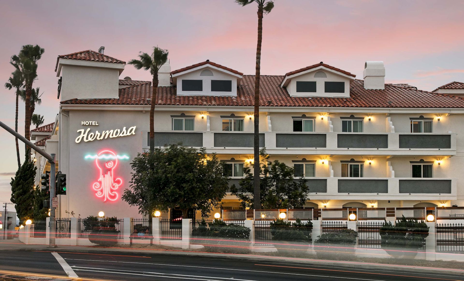 The image shows a hotel with a sign that reads "Hotel Hermosa" and features palm trees and a sunset sky in the background.