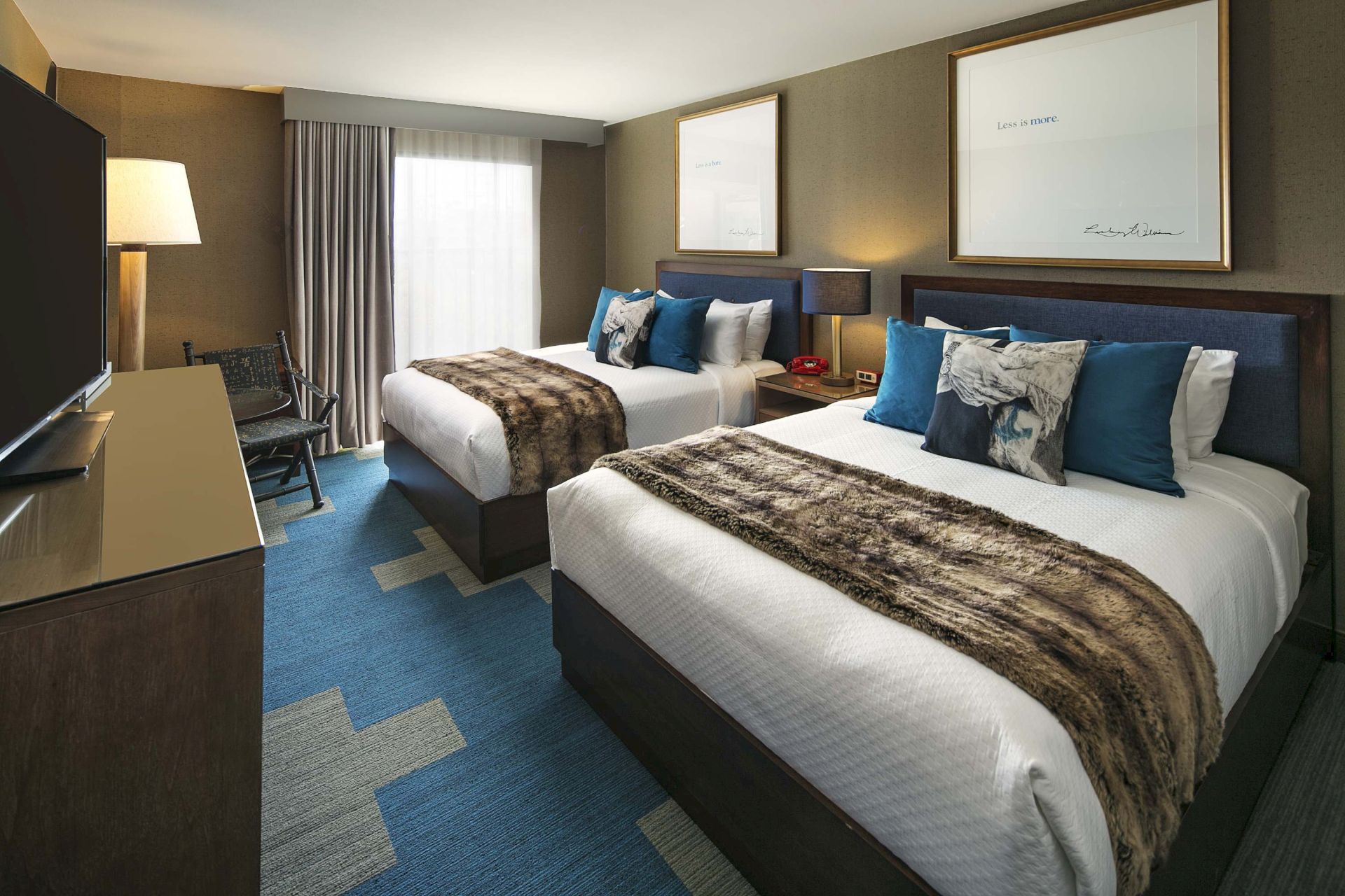 A modern hotel room with two made beds, blue pillows, framed wall art, and curtains near a window.