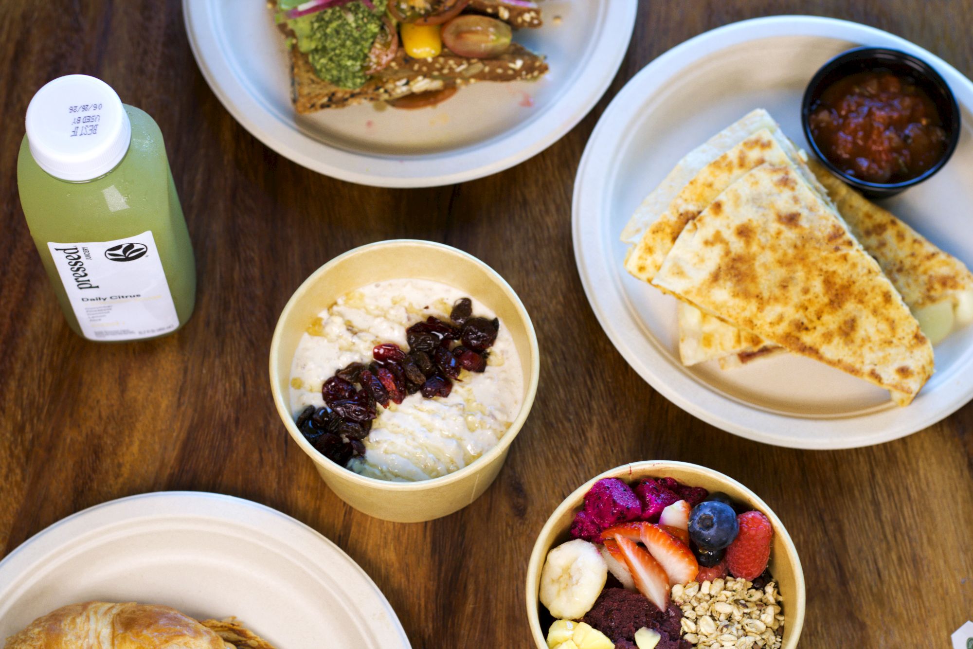 A breakfast spread: quesadilla, yogurt with raisins, fruit bowl, and a green drink bottle on a wooden table.