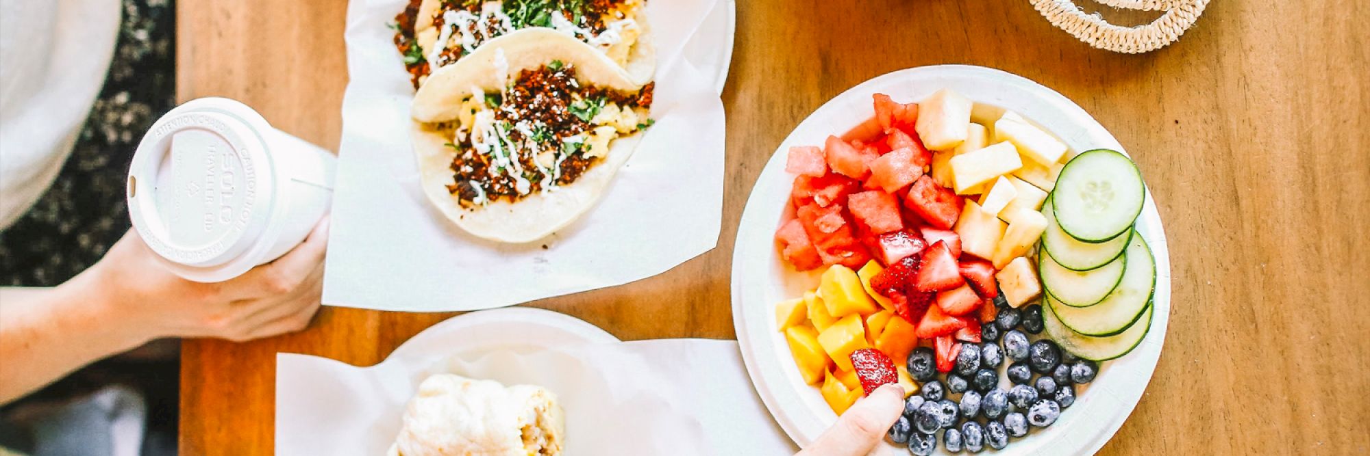 A table with tacos, a plate of mixed fruits and vegetables, a biscuit, and a hand holding a cup of coffee is shown.