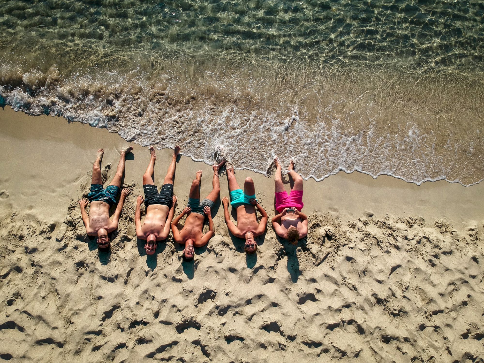 Four people are lying on a sandy beach near the water, each wearing different colored swimwear, with waves gently approaching.