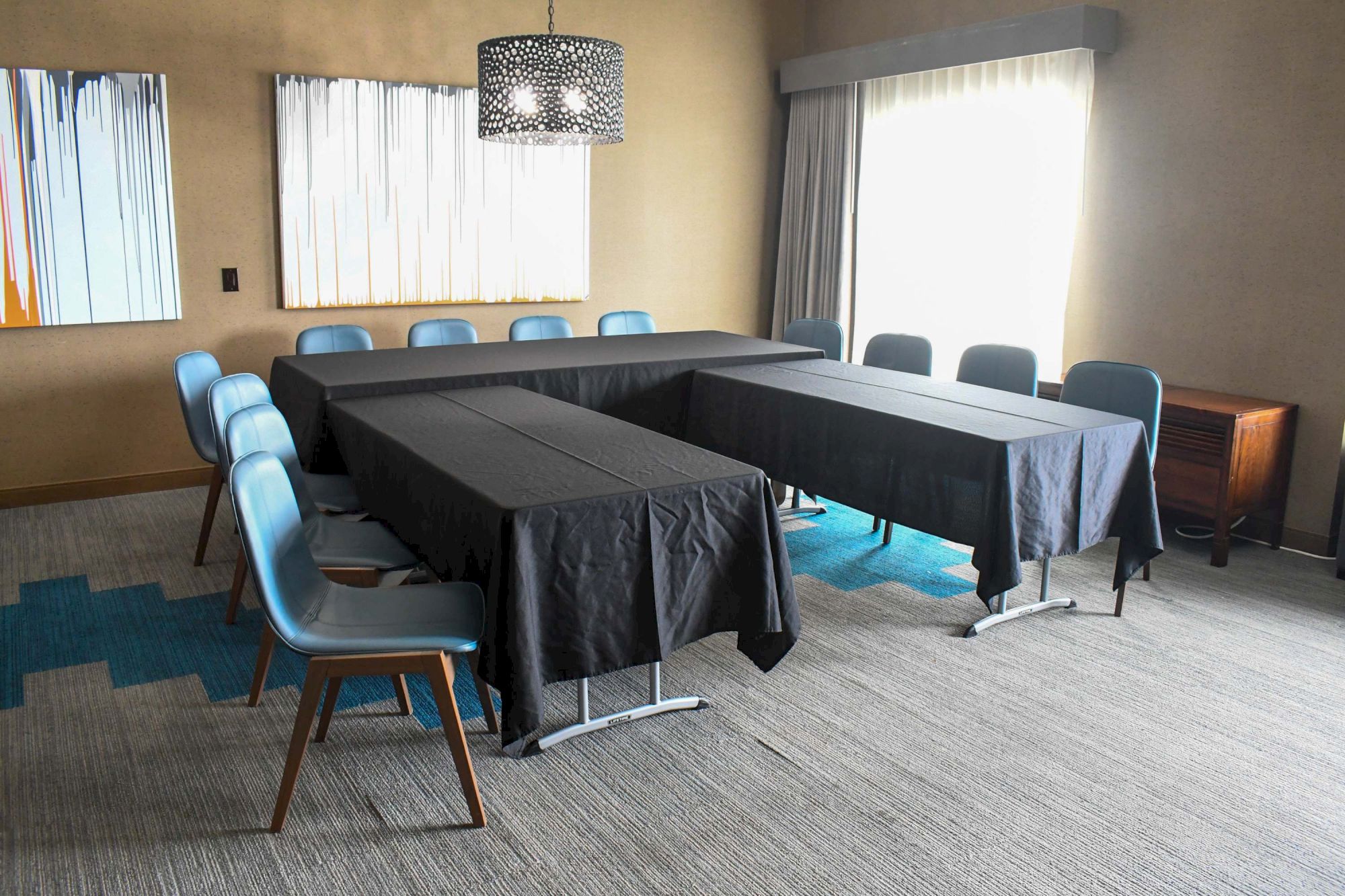 A conference room with U-shaped black table setup, light blue chairs, large window, modern art, and decorated light fixture.