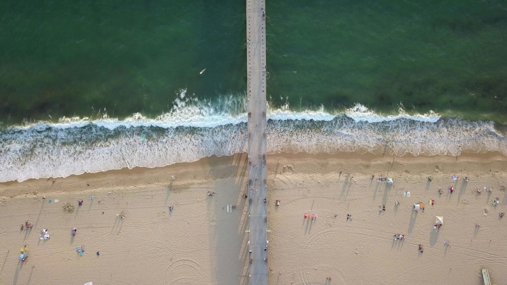 Aerial view of a beach with a pier in the center, dividing the waves and sand, scattered people enjoying the shoreline.