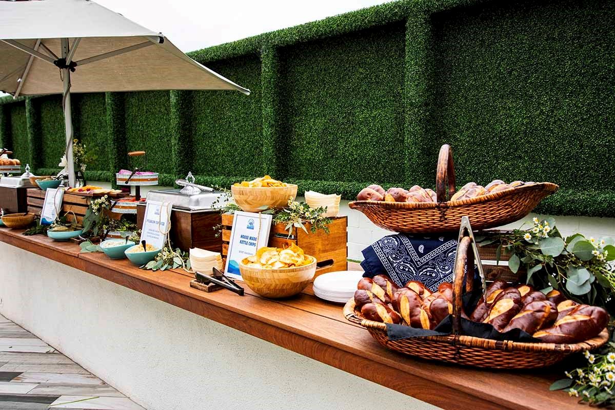 A buffet setup with baskets of bread, bowls of food, and decorative greenery on a wooden counter under an umbrella.