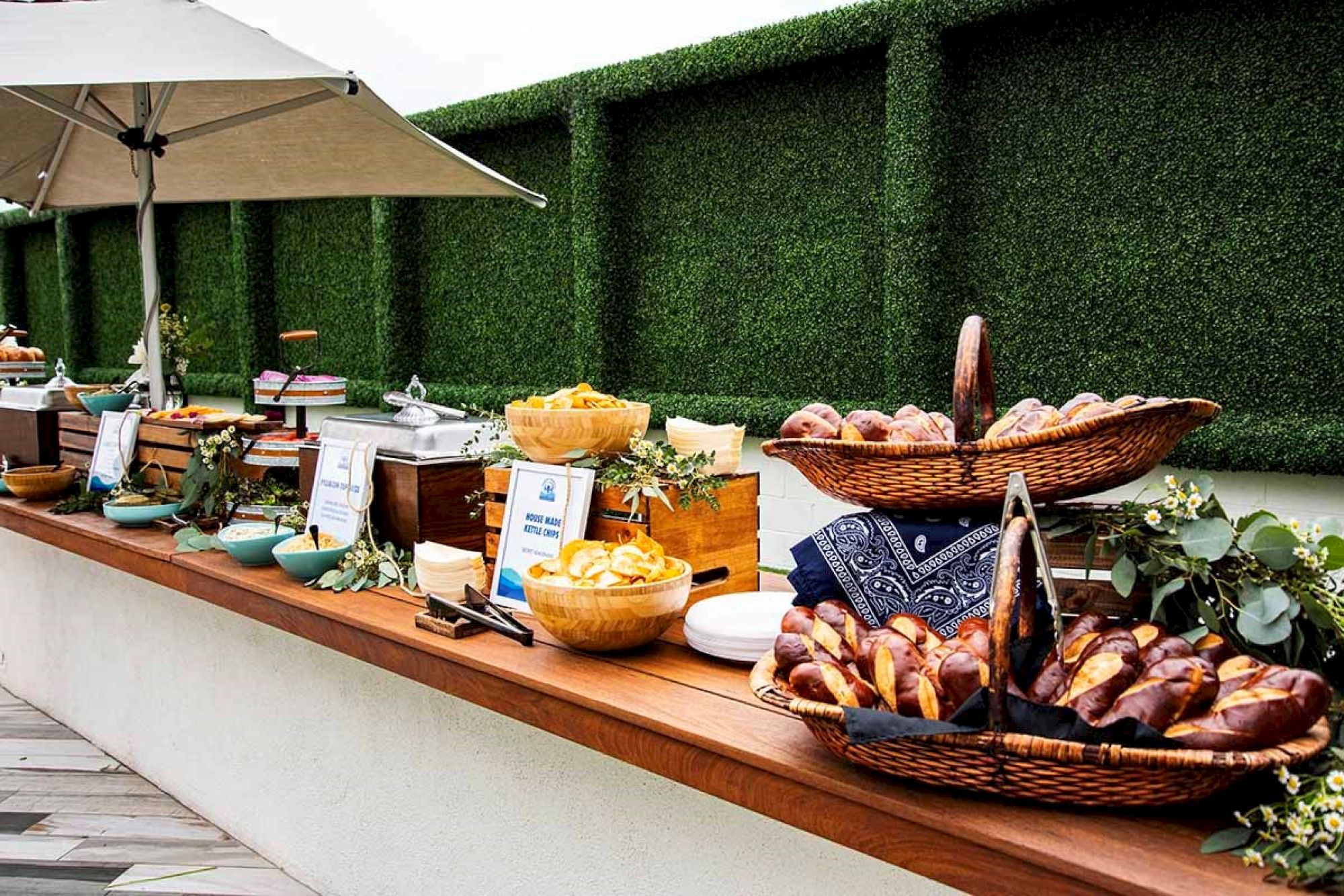 A buffet setup with baskets of bread, bowls of food, and decorative greenery on a wooden counter under an umbrella.