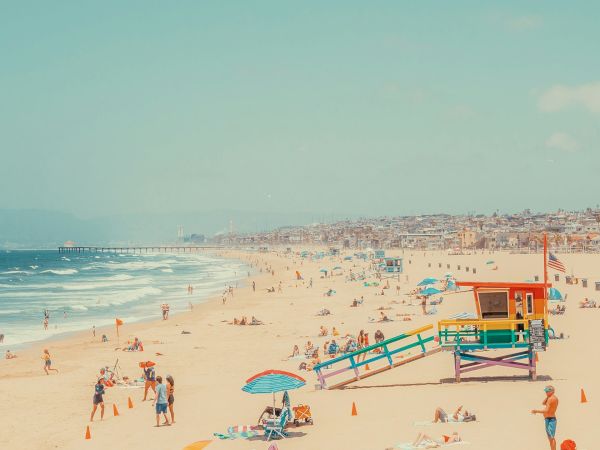 A busy beach scene with people relaxing under umbrellas and a colorful lifeguard station nearby, with waves in the background.