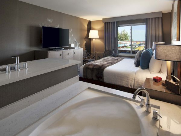 The image shows a modern hotel room with a bed, TV, and a bathroom area with a bathtub.