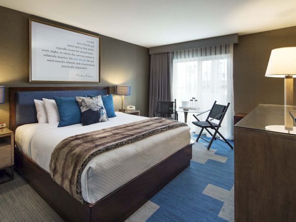 A modern hotel room with a large bed, pillows, a blanket, a wooden dresser, and a window with curtains.