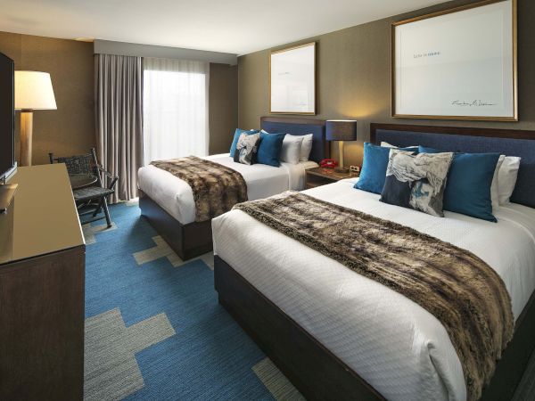 The image shows a hotel room with two double beds, fur throws, decorative pillows, a TV, lamp, and artwork on the walls.