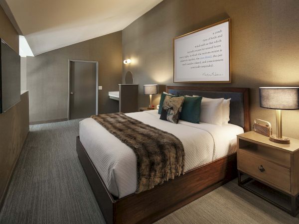 A modern bedroom with a large bed, fluffy pillows, a fur throw, bedside tables, lamps, and wall art, creating a cozy atmosphere.