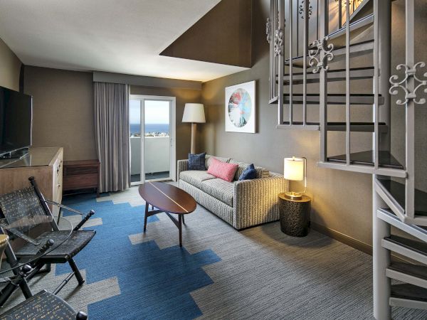 A modern living room features a sofa, coffee table, TV, blue rug, and a staircase, with a view through a glass door to a balcony.