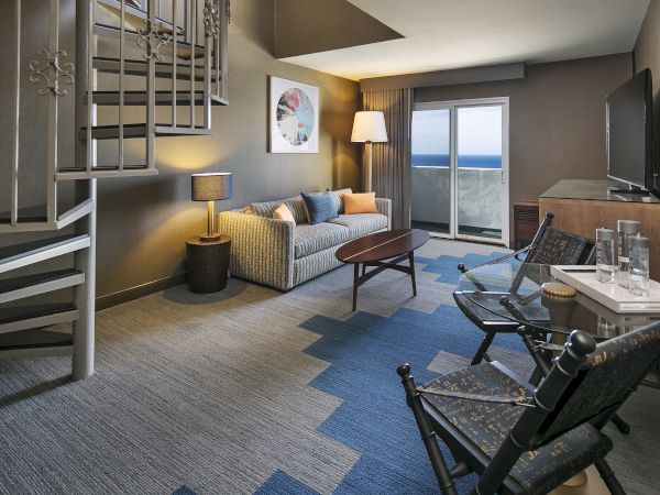 A modern living room with a spiral staircase, sofa, table, and chairs; ocean view through sliding doors, contemporary decor throughout.