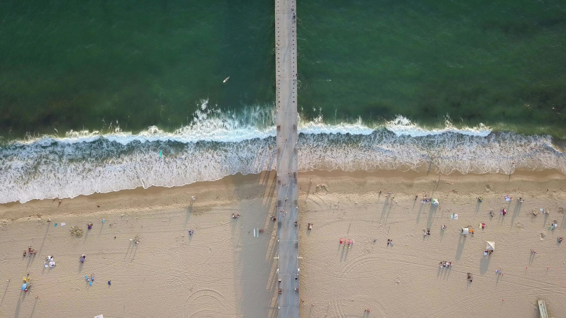 An aerial view of a beach with a pier dividing the ocean and sand, showing people relaxing and enjoying the seaside environment.