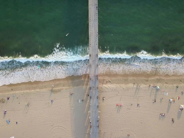 An aerial view of a beach with a pier dividing the ocean and sand, with people scattered along the shore.