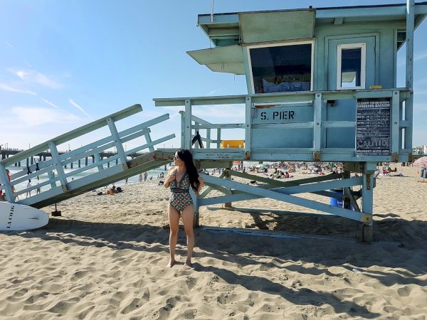 A person in swimwear stands near a light blue lifeguard tower on a sandy beach, with people and surfboard in the background.