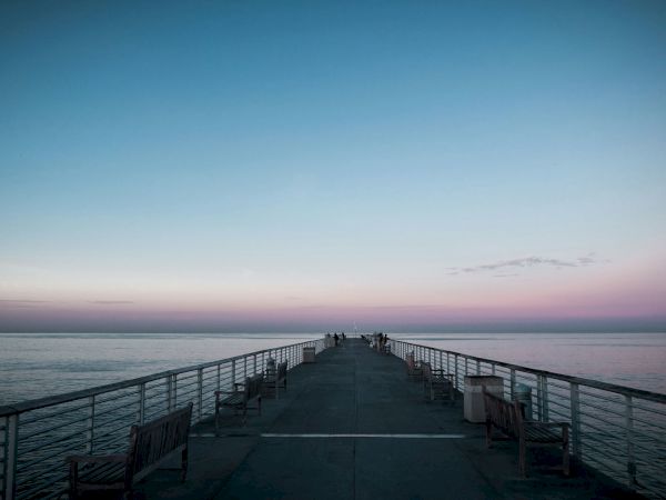 A pier extends into a calm sea at dusk, with benches along its sides and a few people in the distance, under a gradient sky.