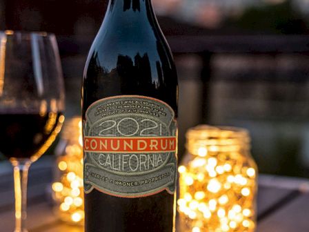 A bottle of Conundrum wine and a glass are on a table, surrounded by glowing string lights in jars, creating a cozy atmosphere.