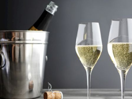 Two glasses filled with champagne are beside an ice bucket holding a champagne bottle, with a cork on the table.