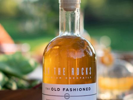 A bottle of "On The Rocks" premium cocktails, labeled "The Old Fashioned," is placed on a table outdoors.