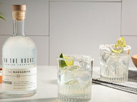 A bottle of "On the Rocks" margarita mix and two margarita glasses with lime wedges, alongside a bowl of limes on a table.