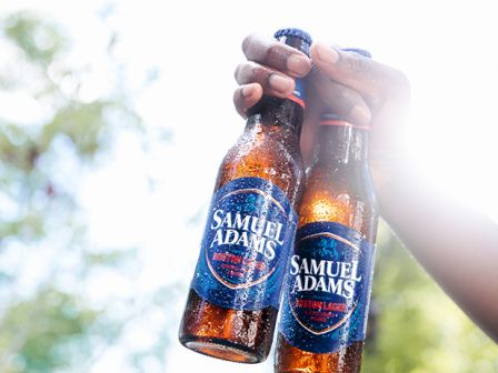 Two hands hold up Samuel Adams beer bottles against a sunny outdoor background.