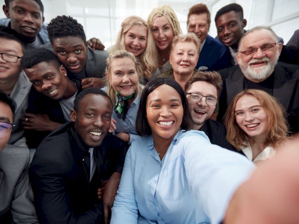 A diverse group of colleagues gathers for a selfie, smiling and posing closely together in a bright office setting. (140 chars)