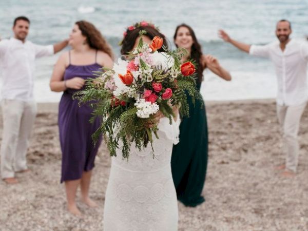 A bride with a bouquet stands on the beach, with four people in the background smiling and reaching out.