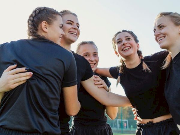 A group of women in black sportswear are smiling and embracing on a sports field, one holding a rugby ball.