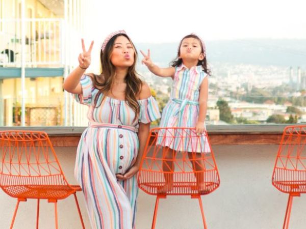 A woman and child pose playfully on a balcony, both wearing matching striped dresses, with three orange chairs beside them.