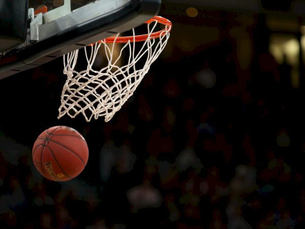 A basketball is about to pass through the hoop during a game in a dimly lit arena.