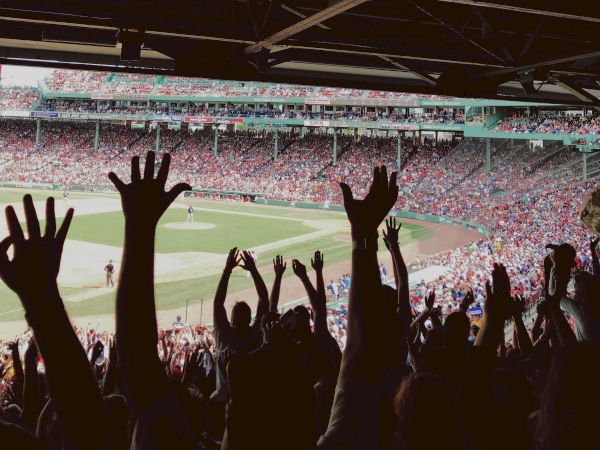 People with raised hands are in a stadium, enjoying a sporting event, with a large field and crowd in the background.