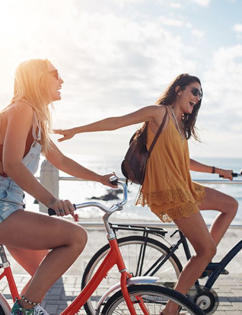 Two people are riding bicycles along a sunny beachside path, smiling and enjoying the day.
