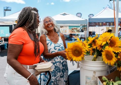 Two women smiling at a farmers' market with sunflowers; one holds a food container, while the other admires the flowers in a bucket.