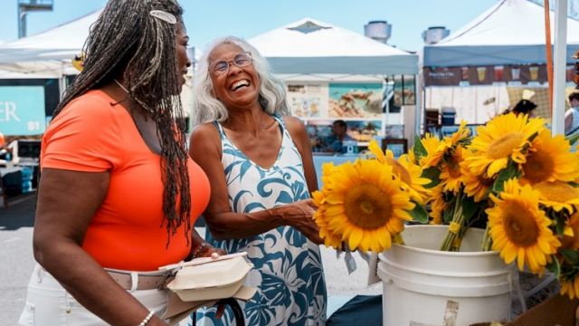 Two women smiling at a farmers' market with sunflowers; one holds a food container, while the other admires the flowers in a bucket.