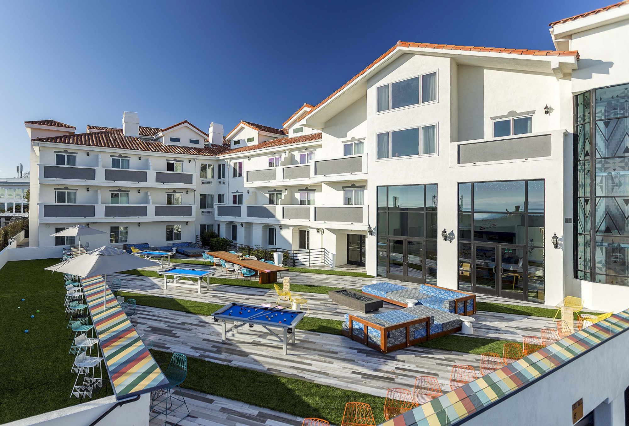 The image shows a modern building with large windows, a patio area with colorful tables, and lounge chairs on a grassy area.