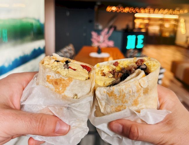 Two hands holding burritos with various fillings inside, set against a modern restaurant interior.