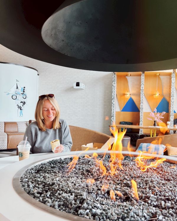 A person sitting by a round fire pit with a drink, inside a stylish modern lounge area with art and geometric decor on the walls.