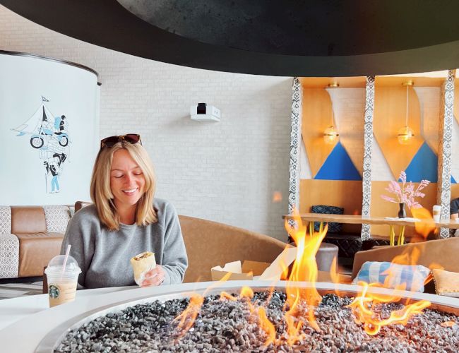 A person sitting by a round fire pit with a drink, inside a stylish modern lounge area with art and geometric decor on the walls.