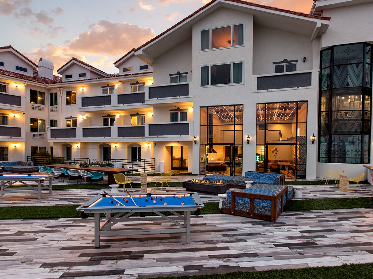 The image shows a modern building with outdoor recreational areas, including tables and games, at sunset.