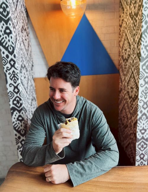 A person is sitting at a table, holding a wrapped sandwich, with geometric and patterned decor in the background.