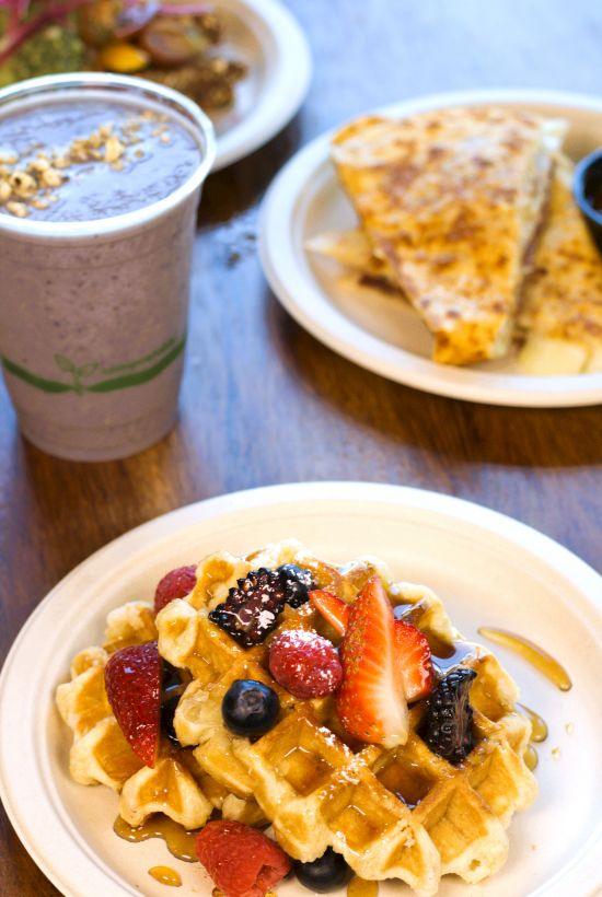 A plate of waffles topped with fresh fruit, a smoothie, and tortilla chips with salsa in the background, all on a wooden table.