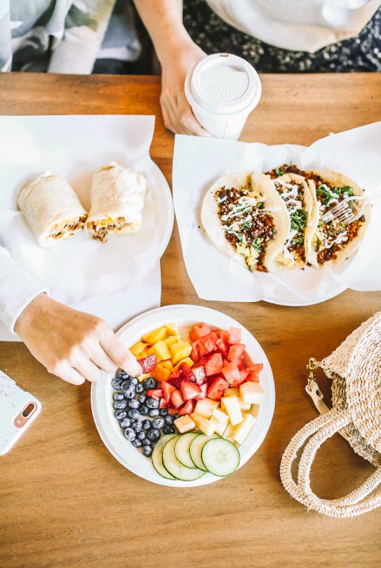 A table with a smoothie bowl, tacos, burrito halves, and a hand holding a drink. A woven bag and phone are also visible on the table.