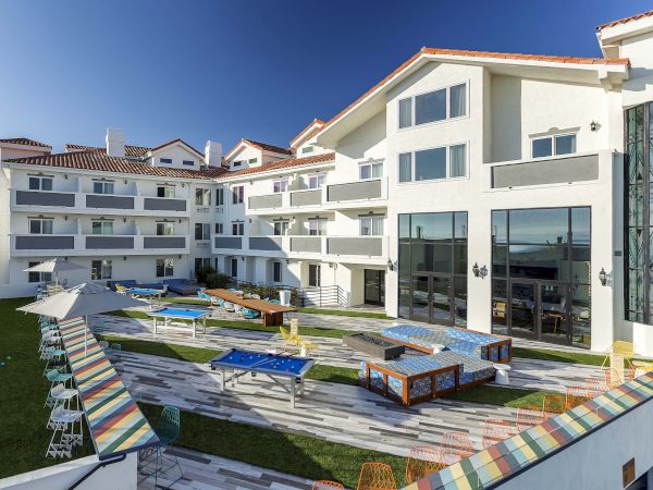 The image shows a modern building with glass panels, a courtyard featuring tables, chairs, and umbrellas under a clear blue sky.