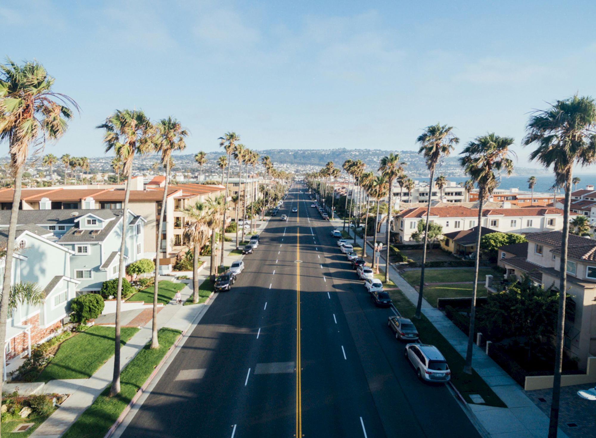 A sunny street lined with palm trees, houses on both sides, and parked cars, leading towards the ocean in the distance.