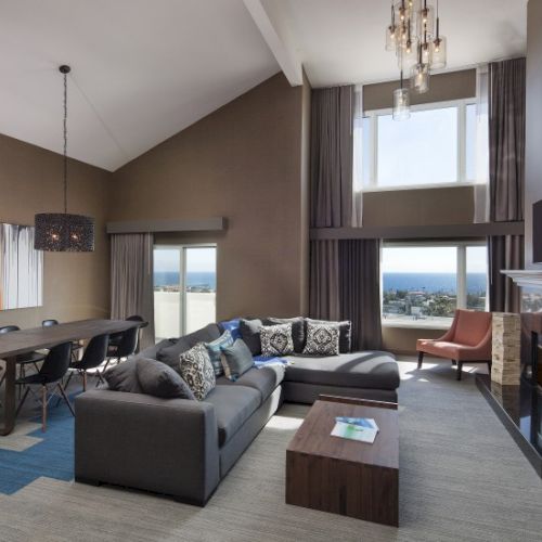 A modern living room with high ceilings, a sectional sofa, dining table, fireplace, and large windows overlooking the sea.