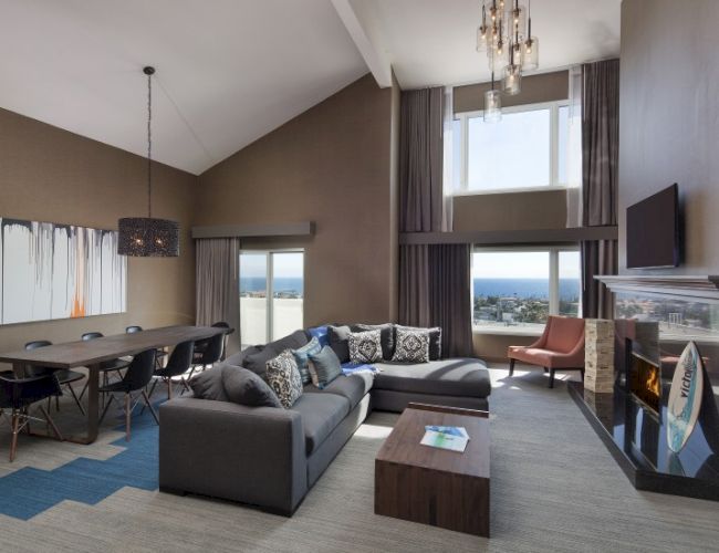 A modern living room with high ceilings, a sectional sofa, dining table, fireplace, and large windows overlooking the sea.