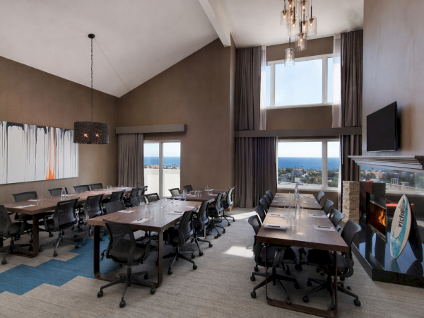 A modern conference room with long tables, black rolling chairs, large windows, a fireplace, a mounted TV, pendant lights, and artwork&mdash;bright view.