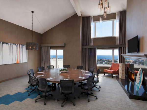 A modern boardroom with a large round table, 12 chairs, tall windows, a fireplace, wall TV, and contemporary art.