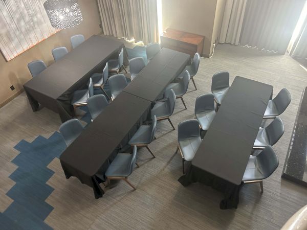 The image shows a meeting room setup with rectangular tables covered in black cloth, surrounded by gray chairs, and some wall decor.